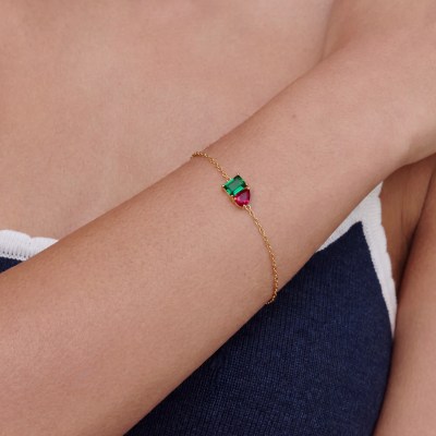 Close-up of a woman's forearm wearing a delicate gold bracelet with a green rectangular gem and a red teardrop gem set in gold.