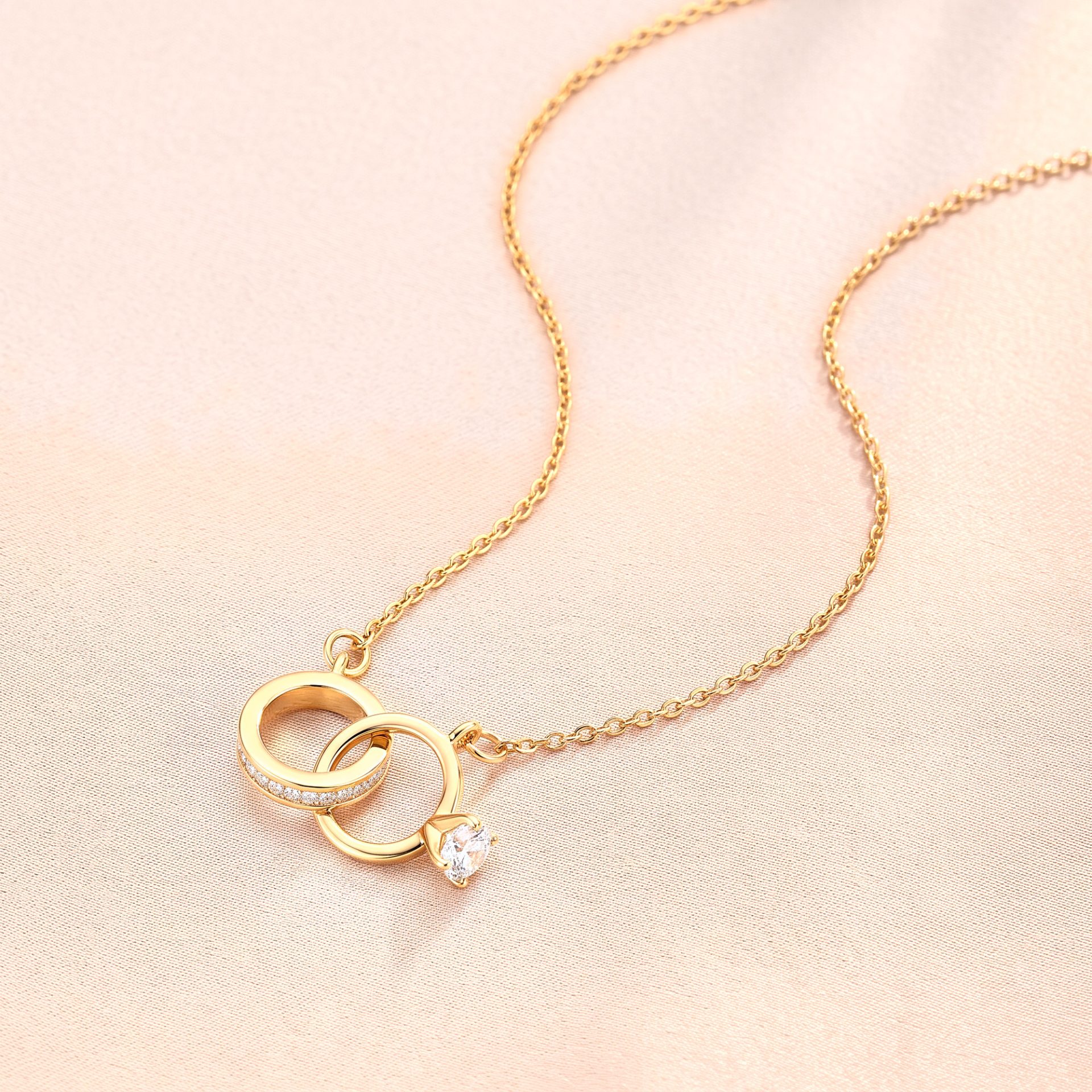Gold chain necklace with interlocking ring pendant and a diamond detail on a soft pink background.