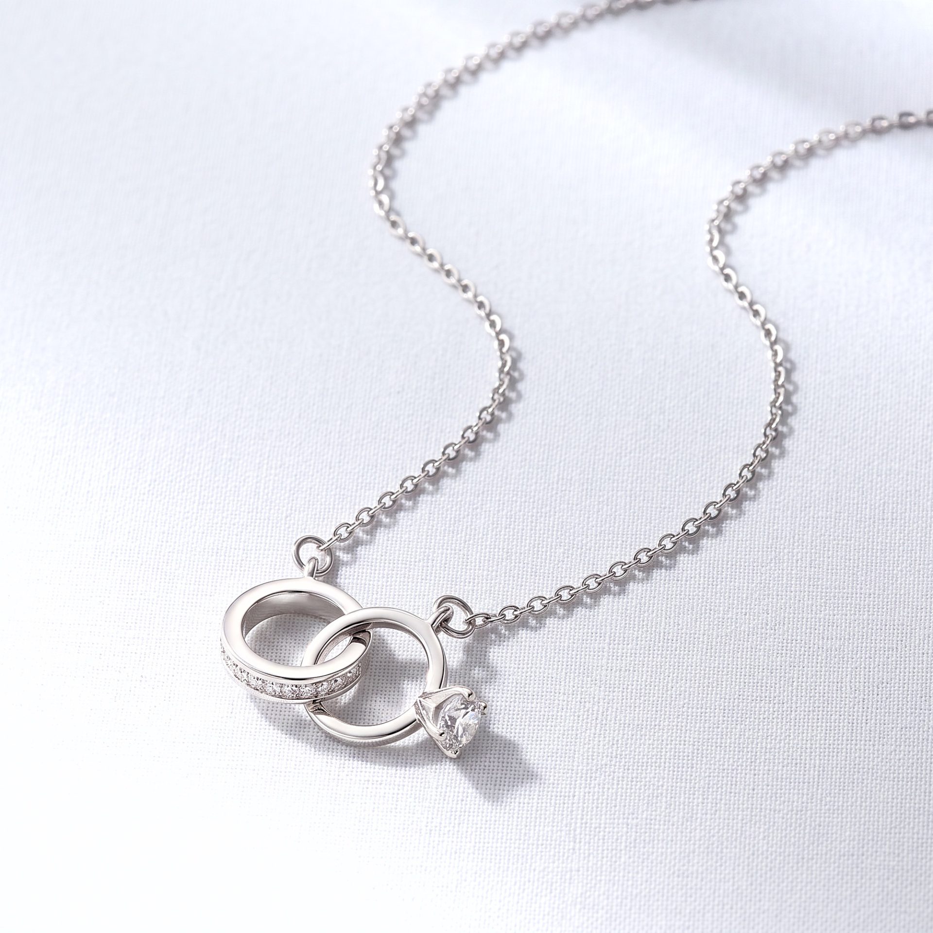 Silver necklace with interlocking ring pendant and a dangling gemstone on a white fabric background.