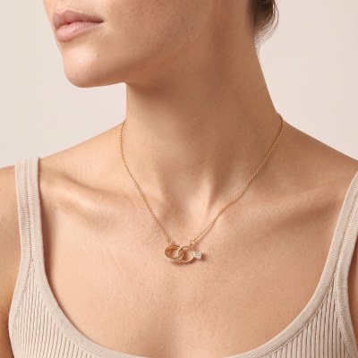 Close-up of a woman's neck wearing a gold chain necklace with two interlocking rings and a dangling gemstone pendant on a beige top.